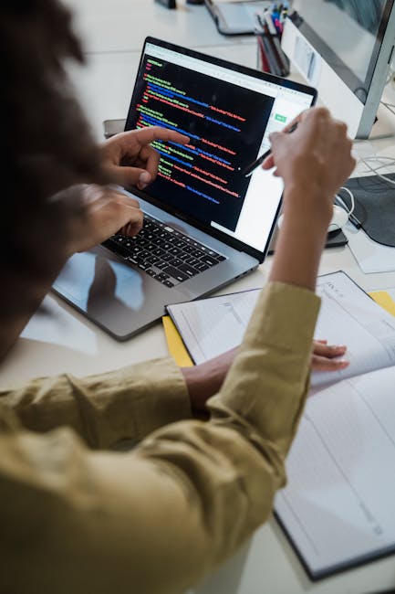 A person working at a desk with a laptop displaying a code editor with multicolored lines of code. The individual is wearing a beige jacket and holding a pen, with a notebook and some documents placed
